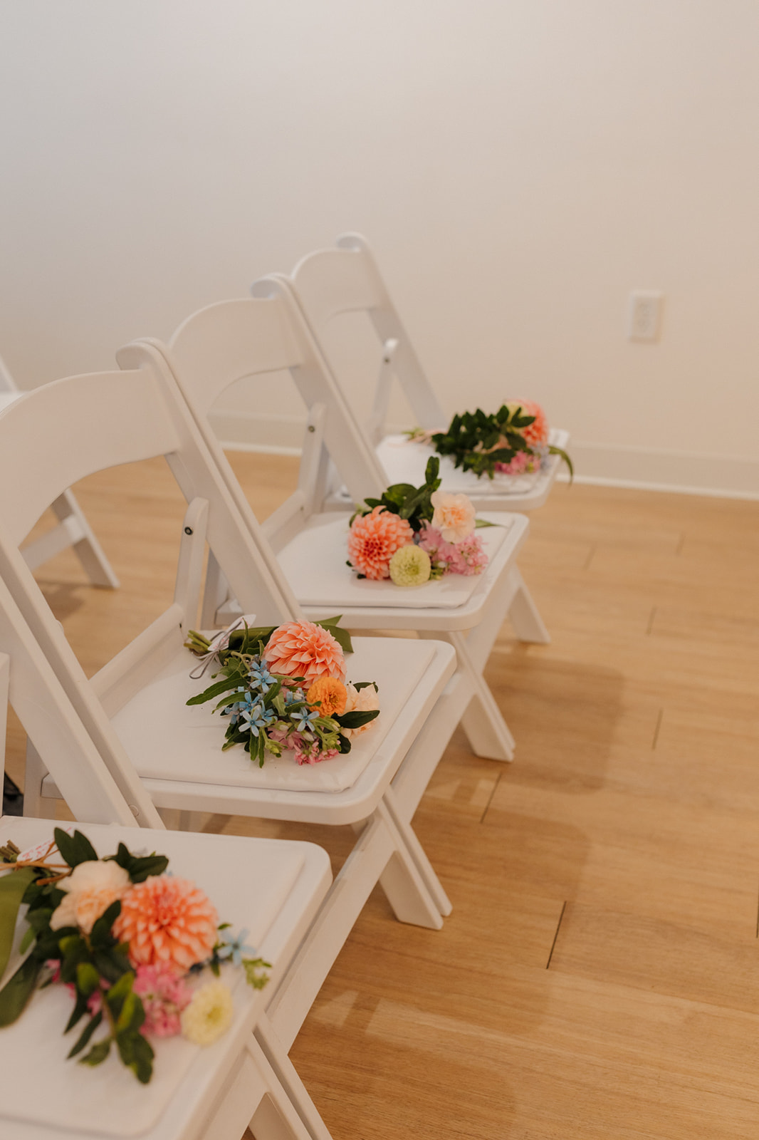 White ceremony chairs lined with fresh floral bundles in peach and blush, waiting for guests to be seated.