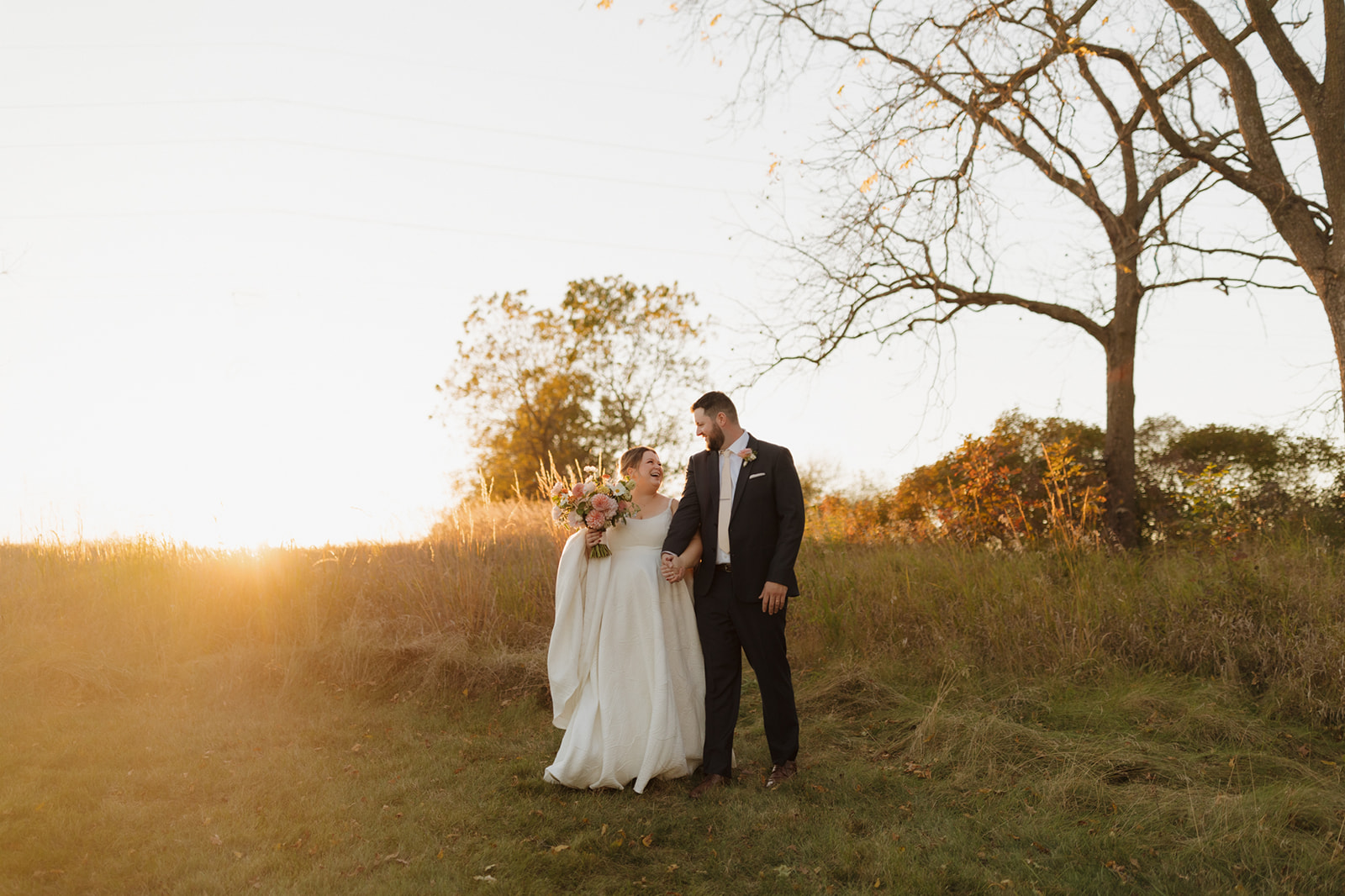 Golden hour stroll through a grassy field as the bride and groom hold hands and share a quiet newlywed moment.