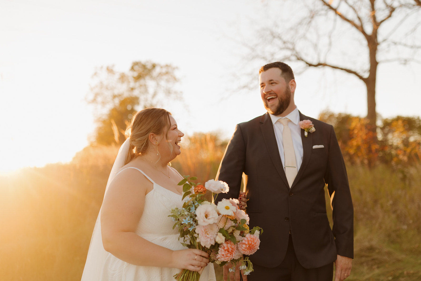 Golden hour laughter between a bride and groom in a field, her bouquet full of pastel blooms and his boutonnière perfectly matching.