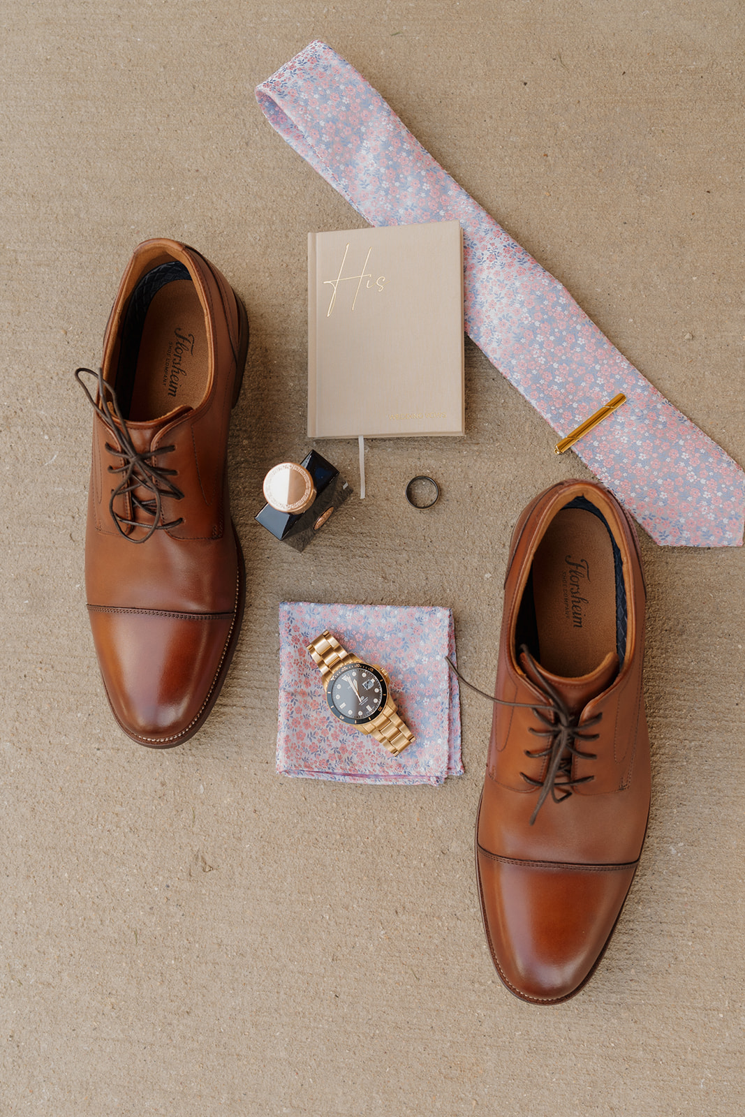Groom’s accessories laid out in a stylish flat lay—classic brown dress shoes, a floral tie, gold watch, and vow book included.