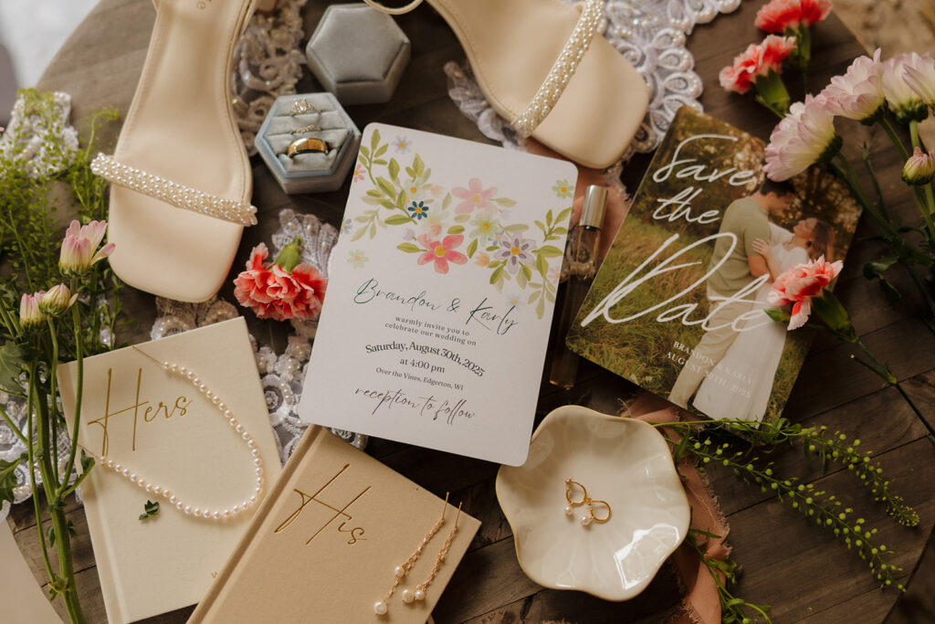 Artful flat lay featuring pearl-detailed heels, wedding rings, florals, and a botanical invitation suite with personalized vow books.