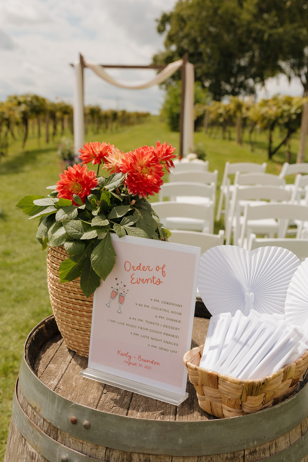 A welcome table with program signage, paper fans, and bright dahlias set atop a wine barrel, greeting guests with charm and style.
