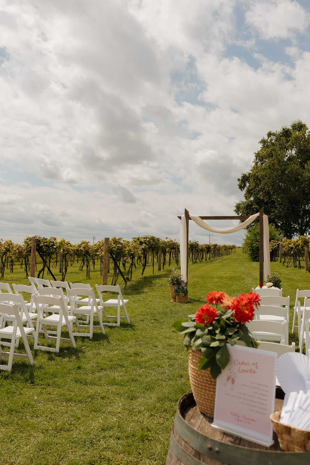 Outdoor wedding ceremony at a vineyard with rows of white chairs, a simple wooden arch, and bright florals under a cloudy sky.