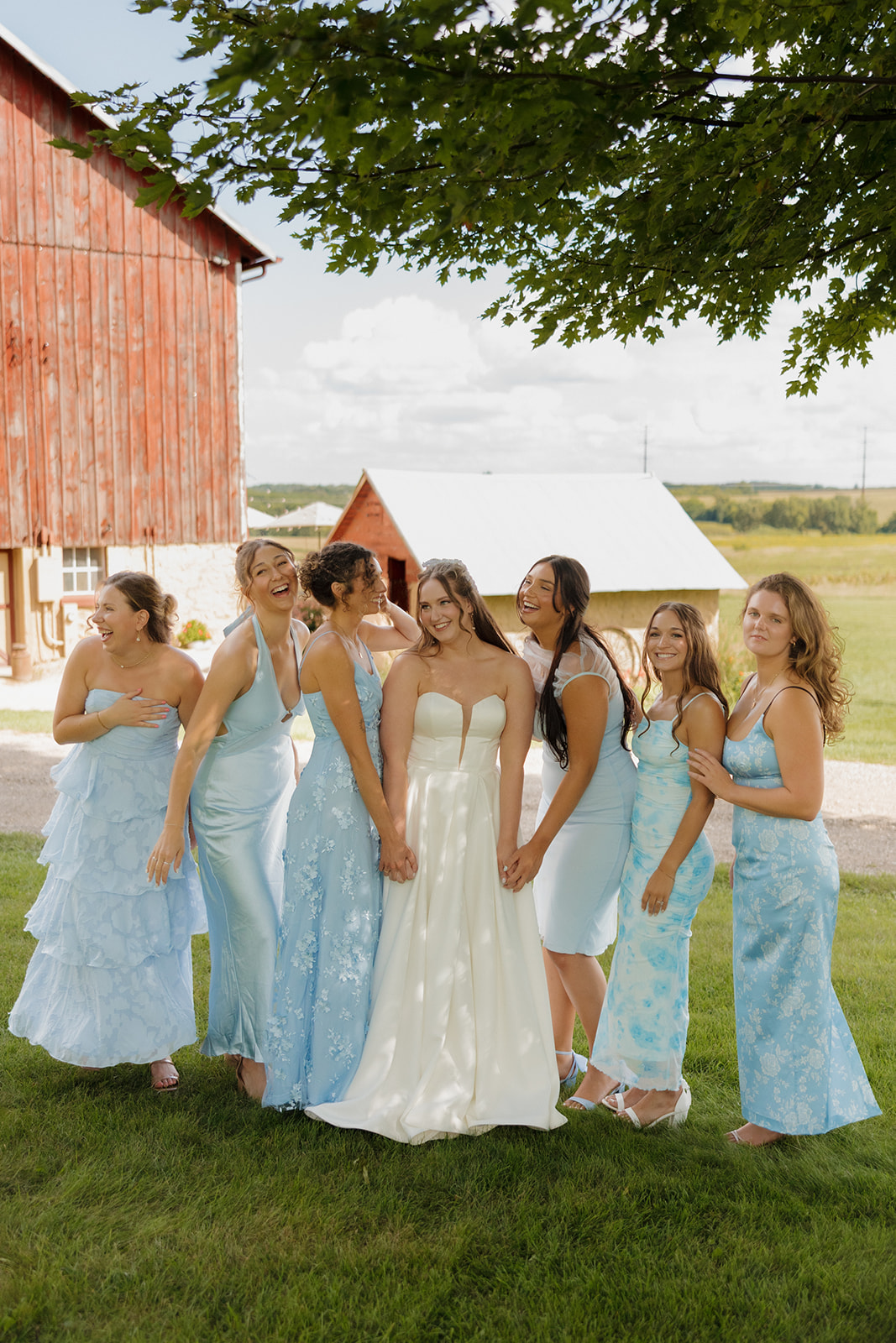 Joyful moment with the bride and her bridesmaids laughing together in soft blue dresses outside a rustic red barn.