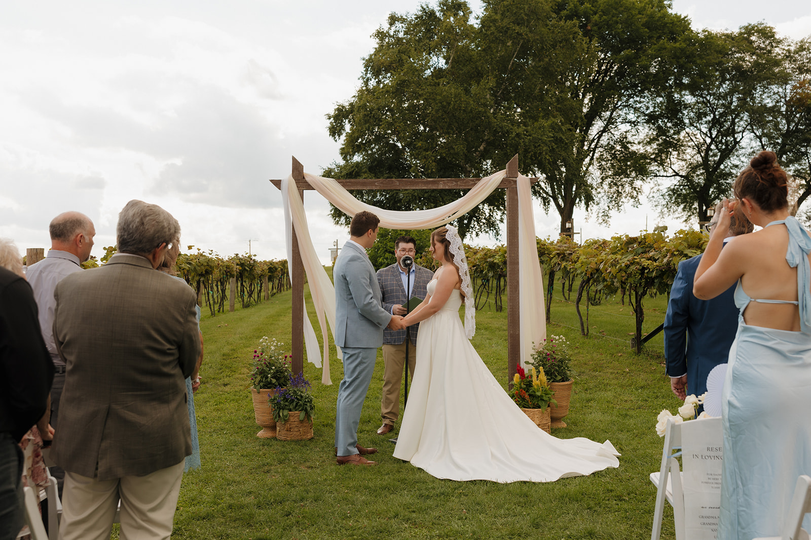 Under a wooden arch draped in fabric, the couple shares vows surrounded by rows of grapevines at a Madison wedding venue.