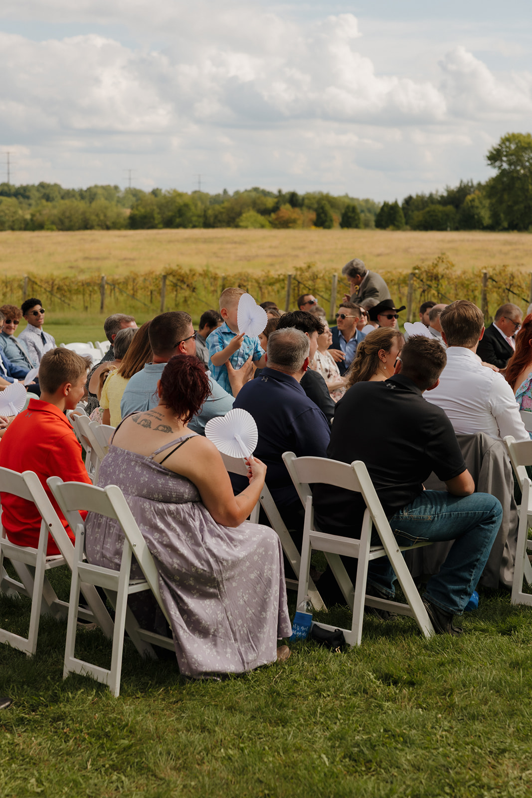 Friends and family settle into white chairs on the lawn, fans in hand, ready for a heartfelt outdoor wedding ceremony.