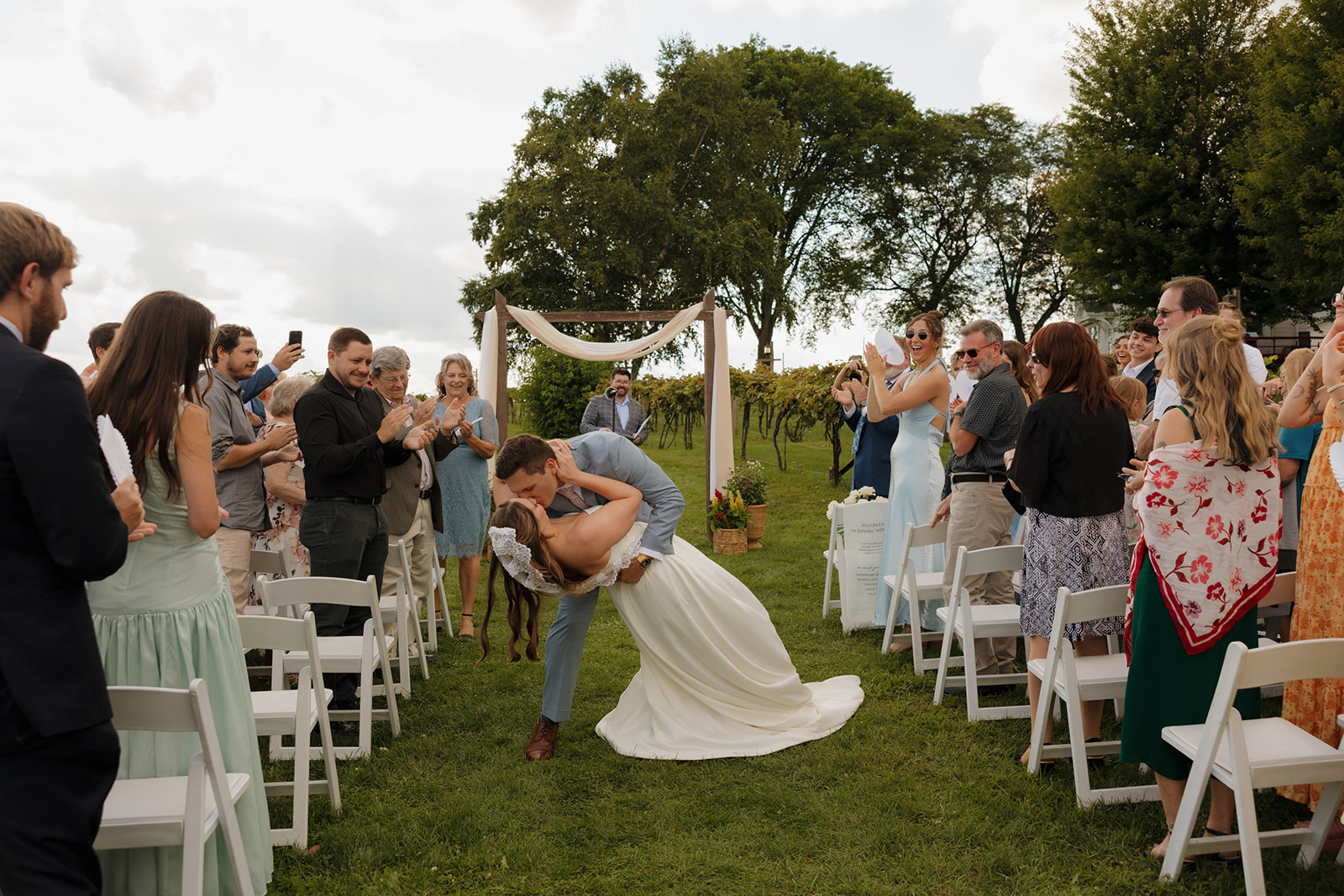 Newlyweds share a dramatic kiss dip at the end of the aisle while guests cheer and clap around them.
