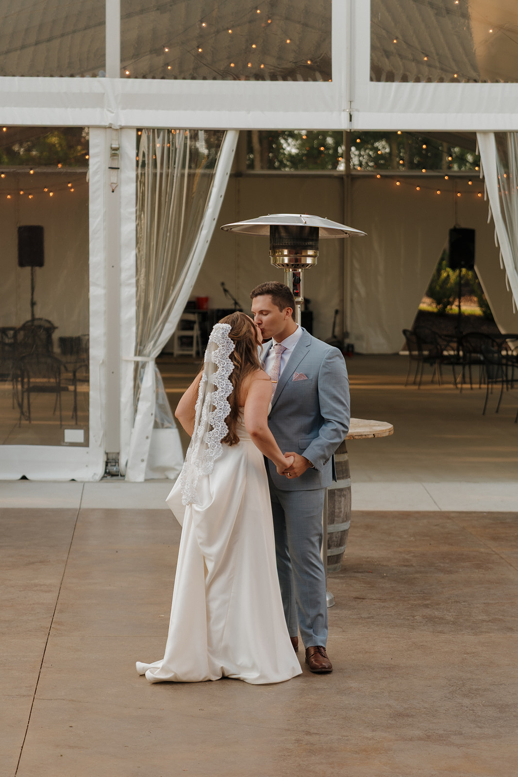 Bride and groom share a quiet moment during their first dance beneath a clear wedding tent with string lights and soft evening light.