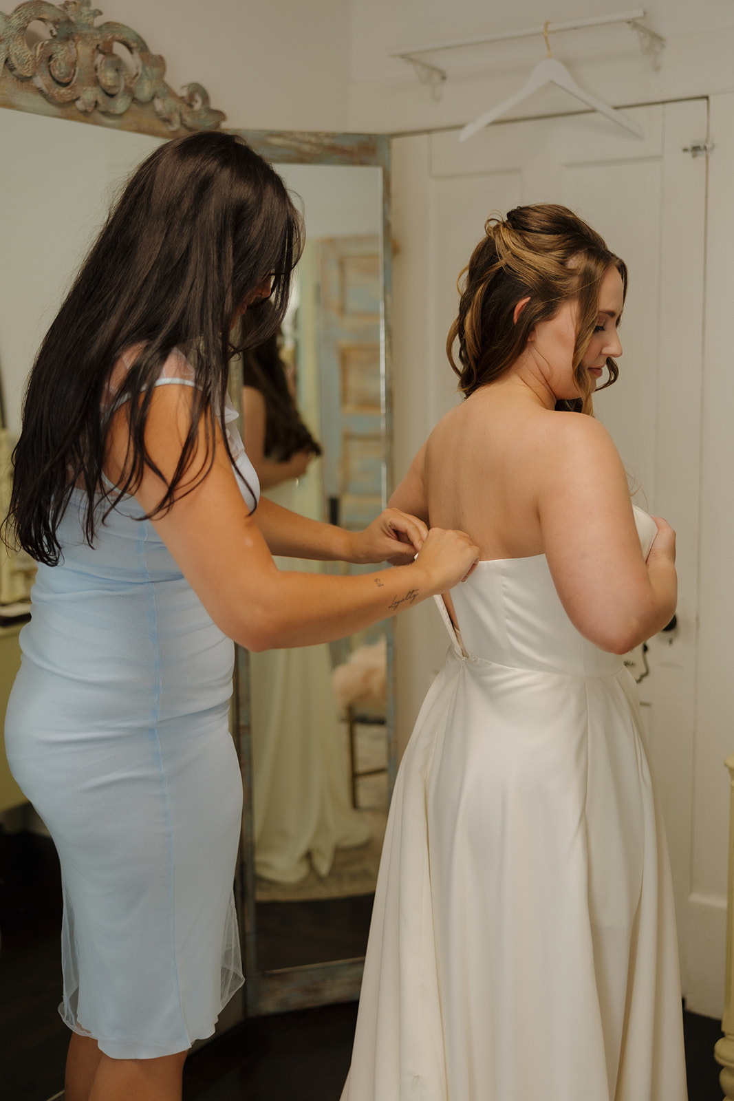 Sweet getting-ready moment as a bridesmaid helps zip up the bride’s gown, surrounded by mirrors and vintage charm.