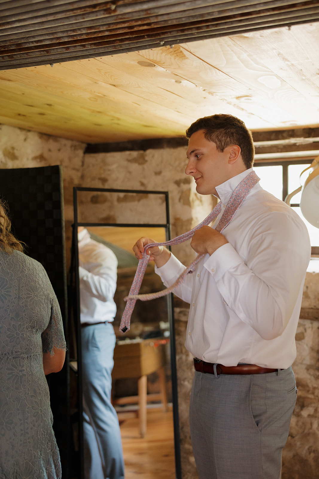 A candid side view of the groom tying his pink tie, sunlight streaming through the stone-walled groom’s suite.