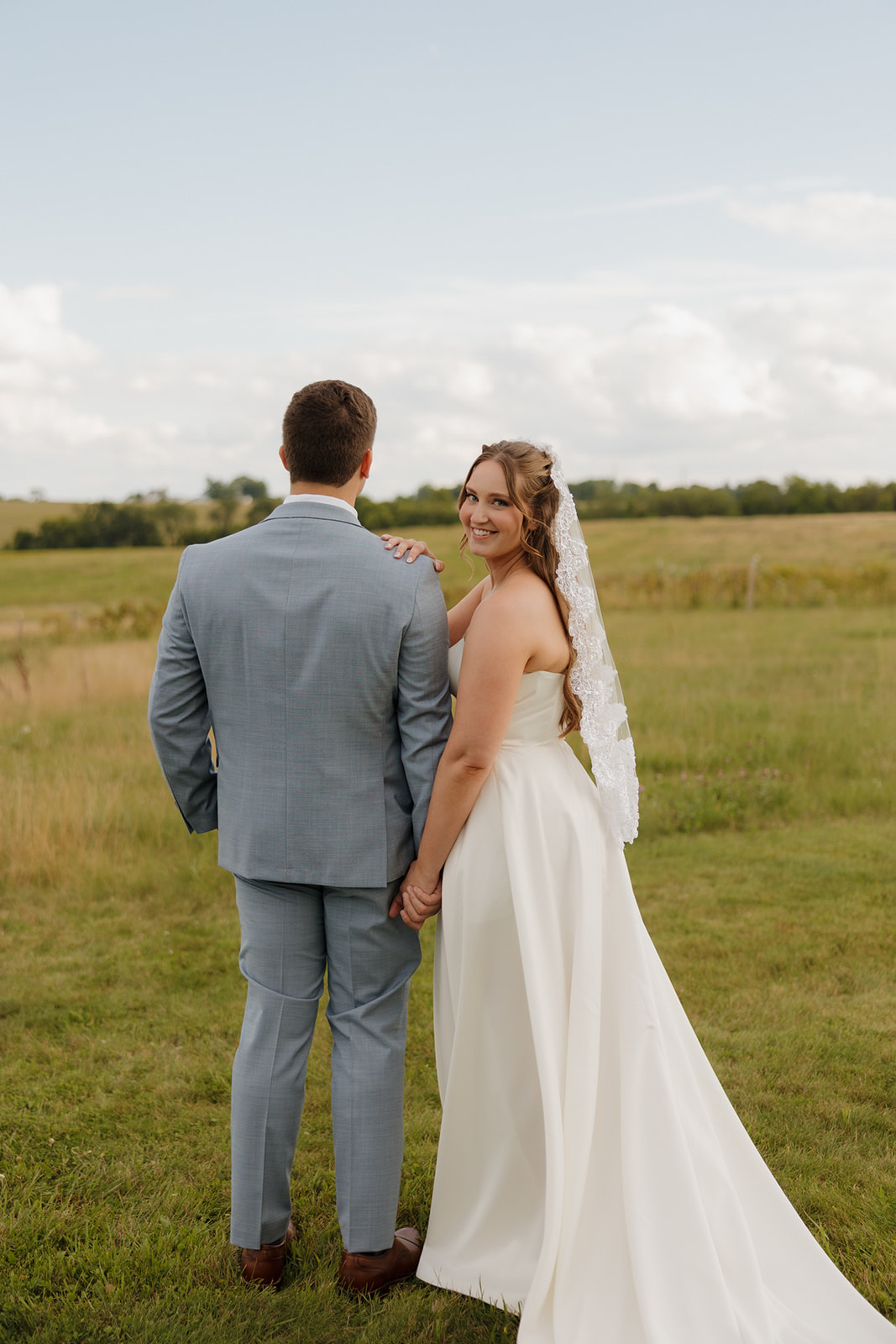 The bride flashes a glowing smile over her shoulder while holding hands with her groom in the wide Wisconsin countryside.