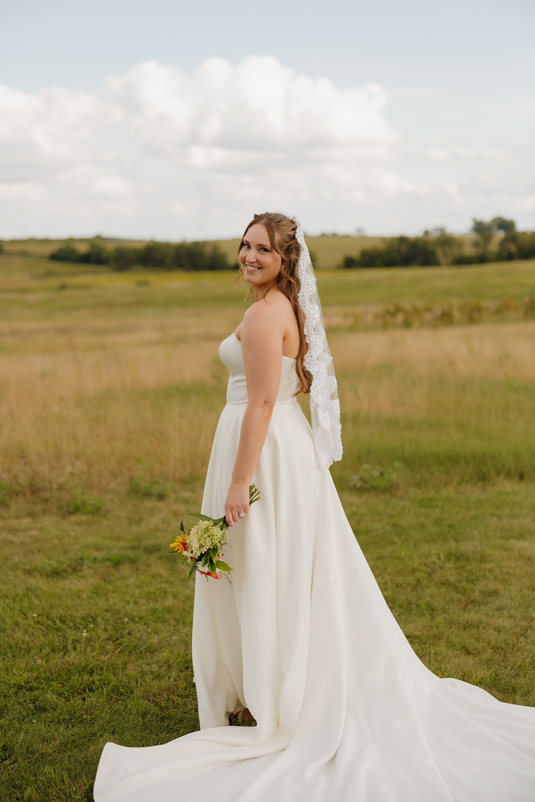 Timeless bridal portrait in a natural field, gown flowing behind and bouquet in hand with a joyful smile.