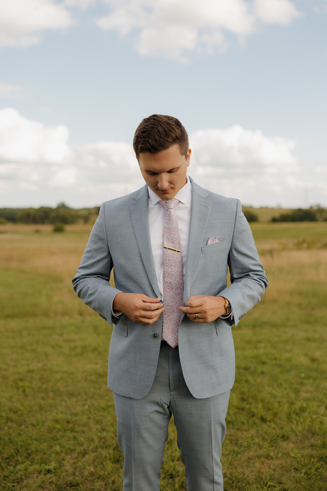 Groom preps for portraits in a field, buttoning his jacket under blue skies and surrounded by gentle hills.