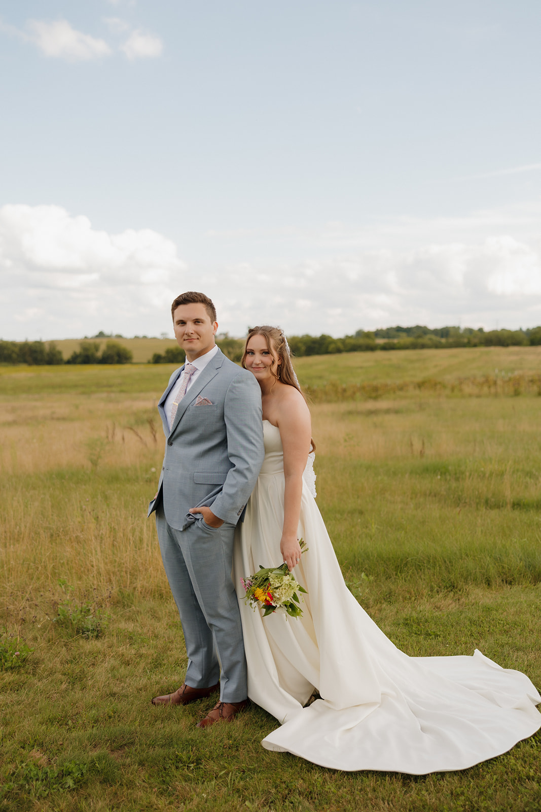The couple stands together in a wide field, the bride holding a bouquet and her gown trailing softly behind.