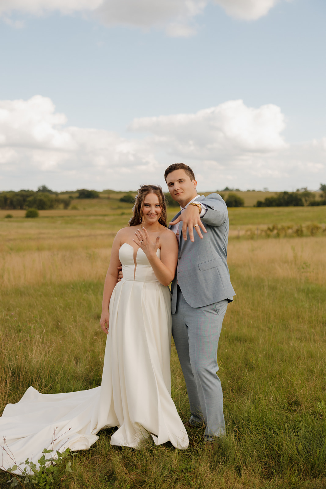 Playful newlywed portrait as the couple flashes their rings and smiles at the camera in a sunlit field.