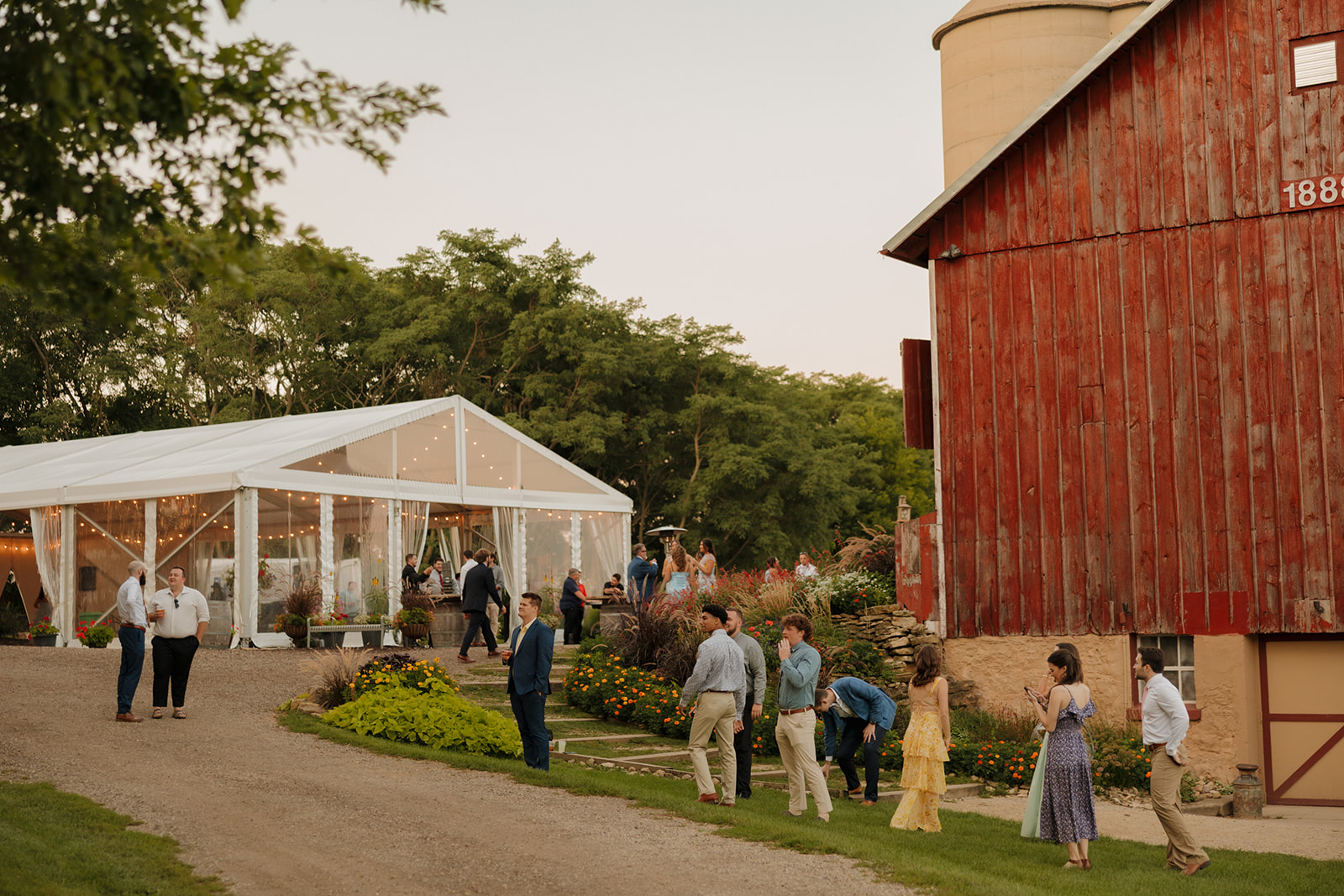Guests gather between a rustic red barn and a glowing clear tent at one of the most inviting Madison wedding venues.