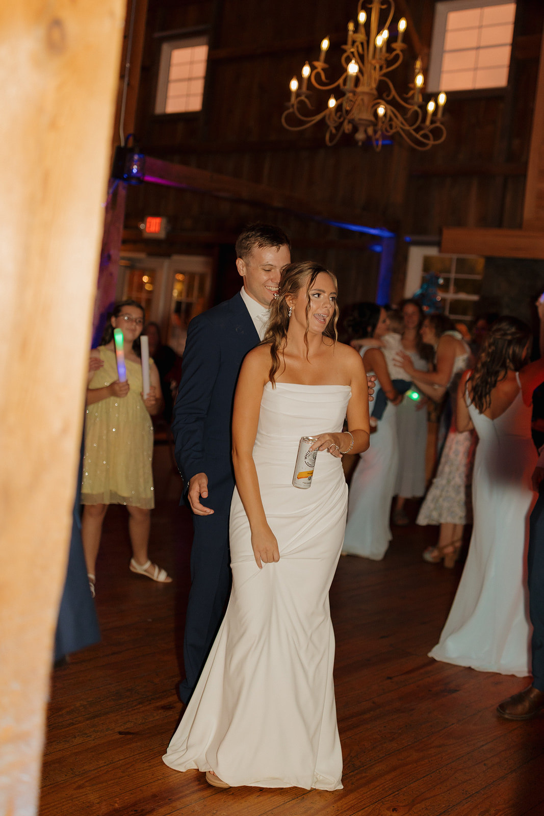 The couple dances together in a warmly lit barn, surrounded by guests and soft chandelier glow, capturing that just-married bliss.