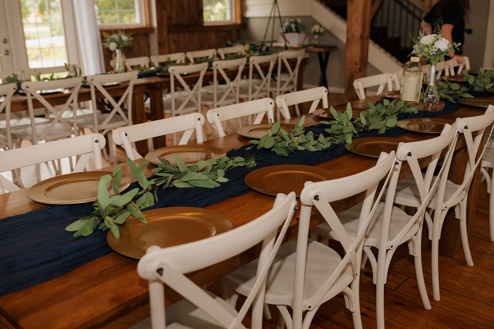 Long wooden reception tables styled with navy runners, greenery garlands, and gold chargers—rustic elegance meets timeless detail in this Wisconsin barn venue.