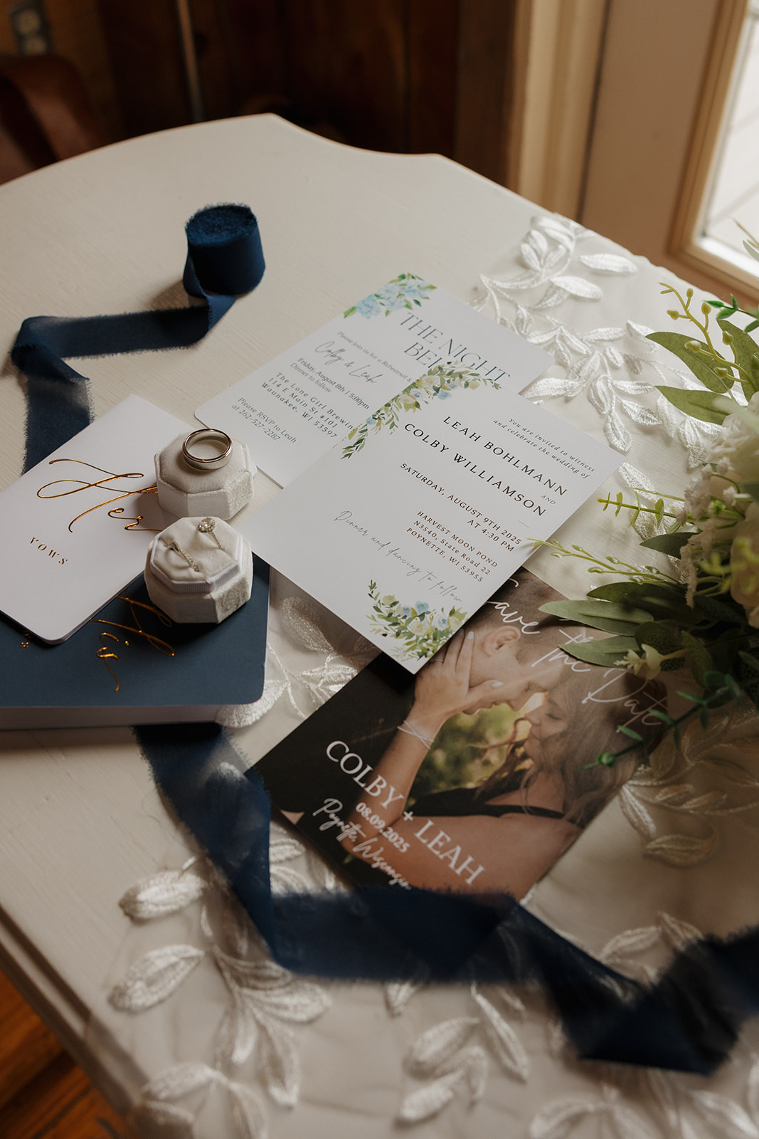 A styled flat lay featuring wedding invitations, vow books, rings, and a navy ribbon on a lace-covered table—captured with detail by a wedding photographer in Wisconsin.