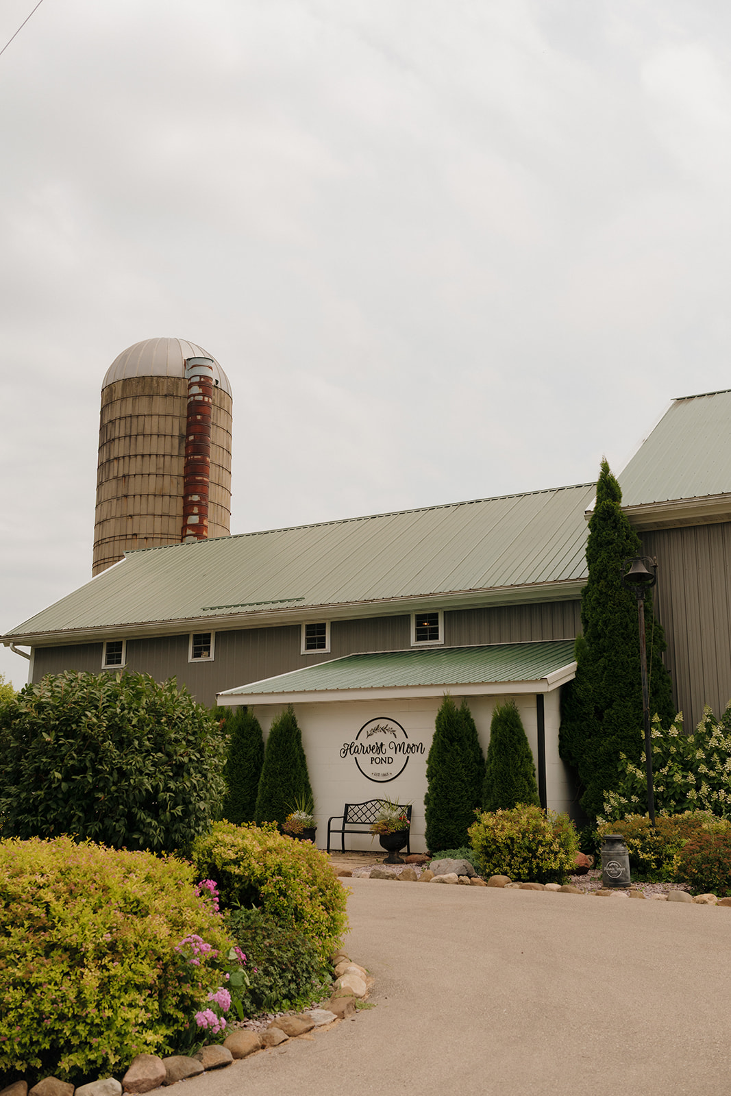 A wide view of the Harvest Moon Pond barn with a green roof, silo, and lush landscaping, welcoming guests to this rustic Wisconsin wedding venue.