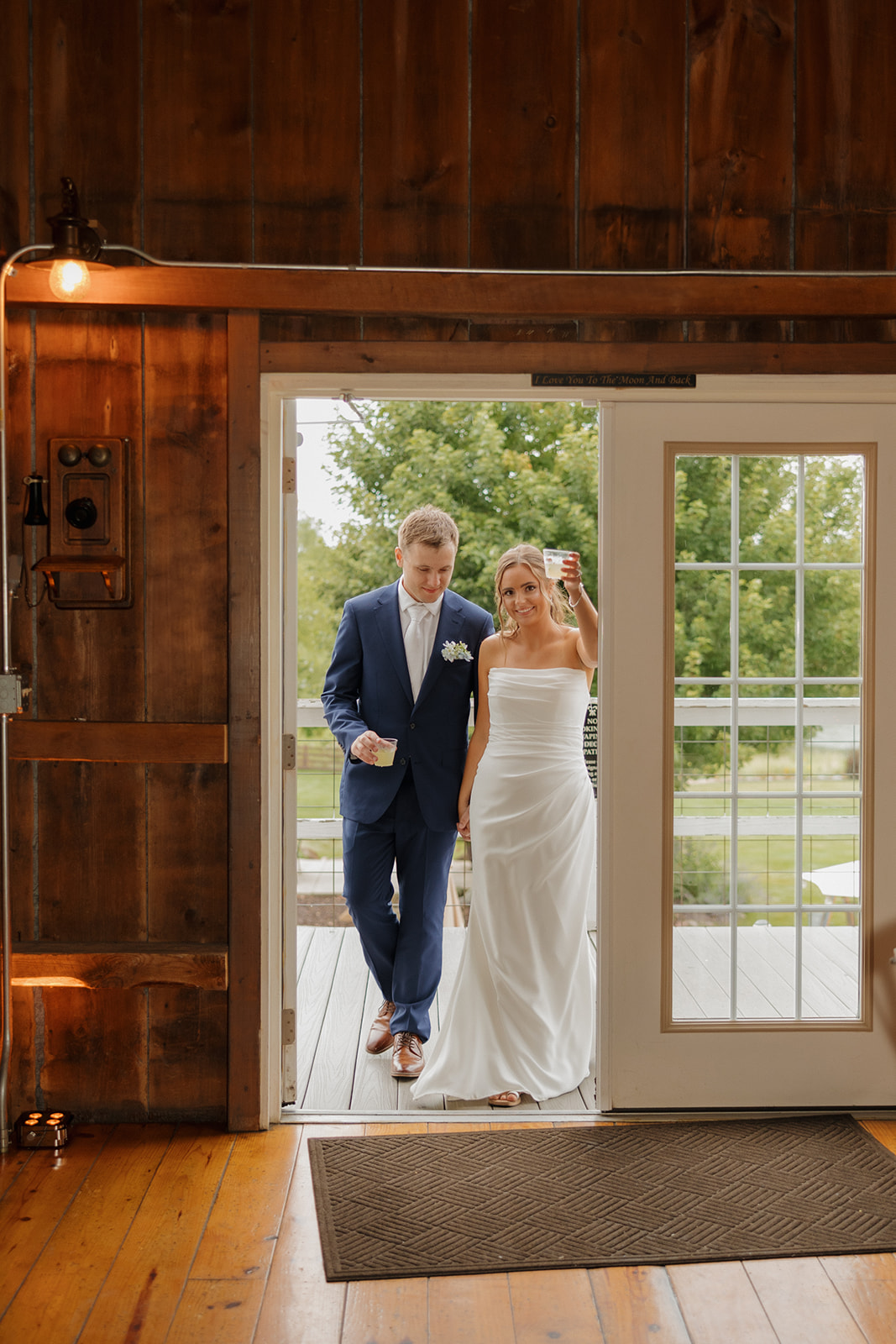 The bride and groom make their entrance hand-in-hand through white barn doors, drinks in hand and smiles ready, into their rustic-chic reception.