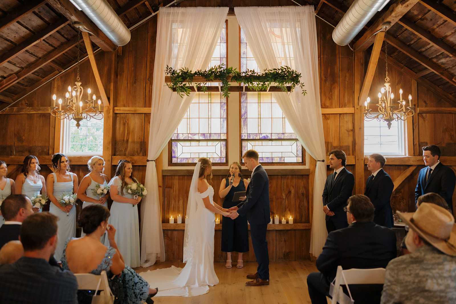 The couple holds hands in front of their officiant, framed by stained glass, candles, and wooden walls—an intimate barn ceremony filled with light and love.