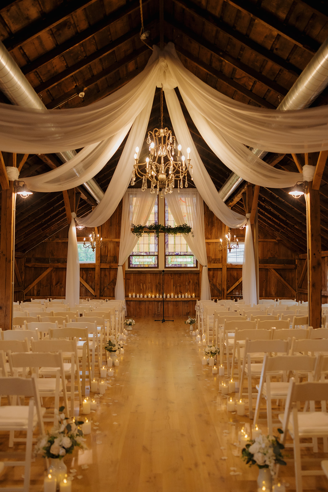 A romantic barn ceremony setup with white chairs, chandeliers, soft draping, and candlelit aisle, designed for a cozy and elegant wedding day.