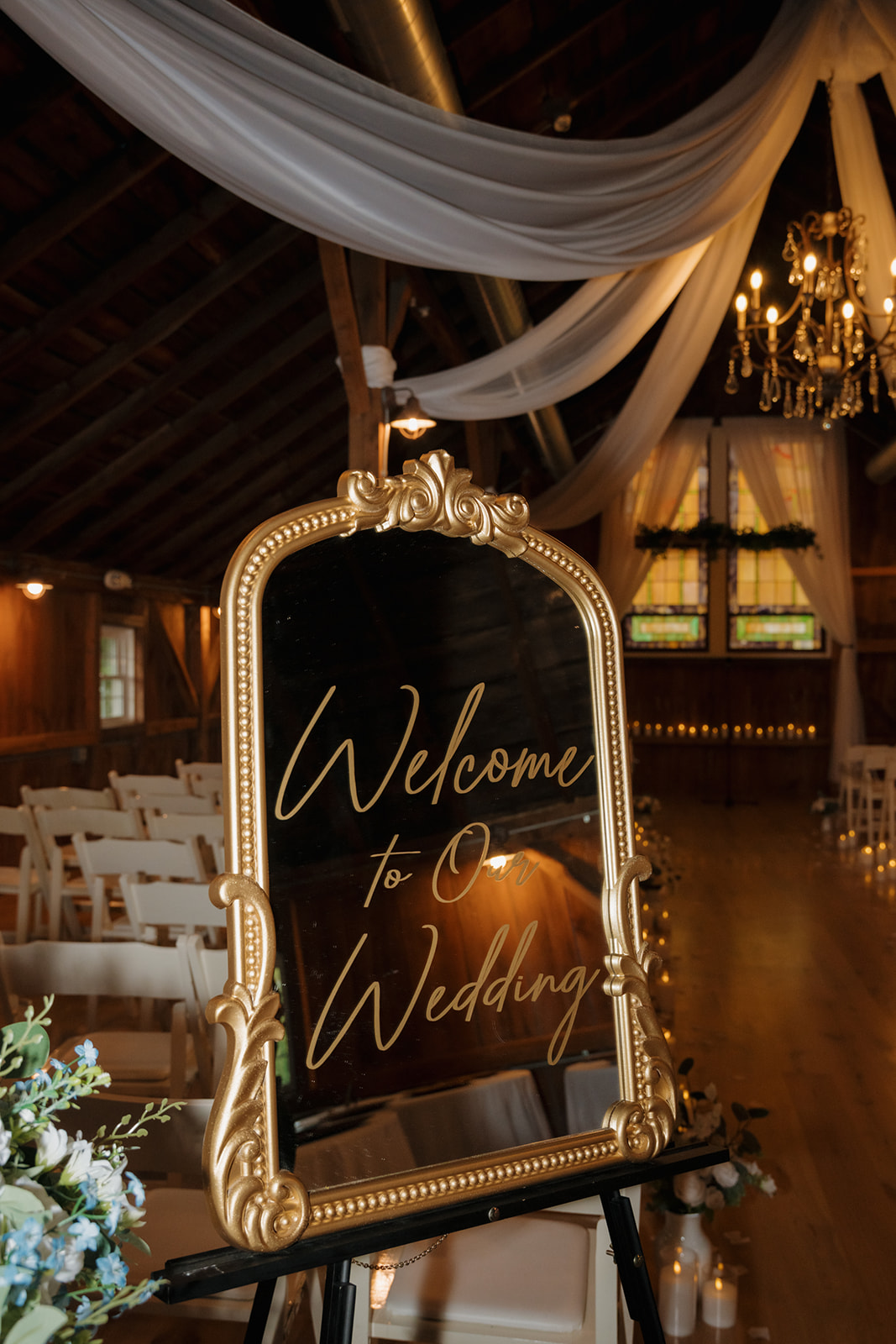 A gold-framed mirror with “Welcome to Our Wedding” in elegant script greets guests inside a candlelit barn ceremony space draped with white fabric and chandeliers.