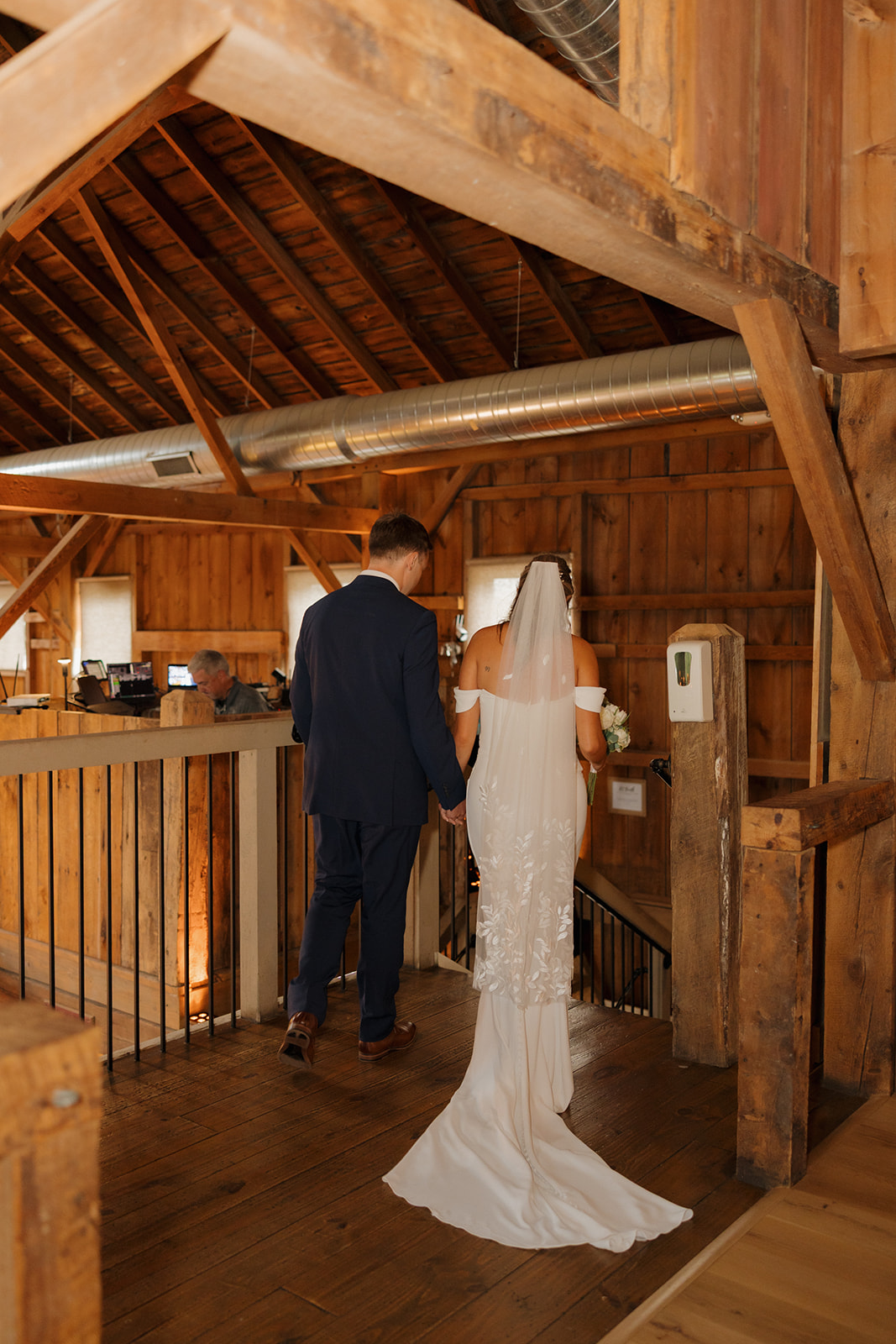 Newlyweds hold hands as they descend the wooden staircase inside a warmly lit barn, surrounded by rich timber beams and rustic charm.