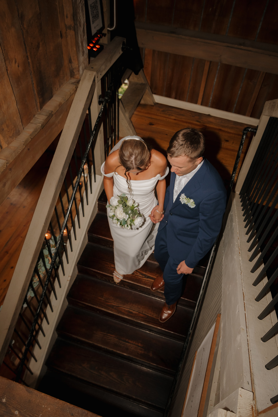 A top-down view of the couple ascending rustic wooden stairs inside a barn, hand-in-hand with bouquet in tow, showcasing the details of their attire.
