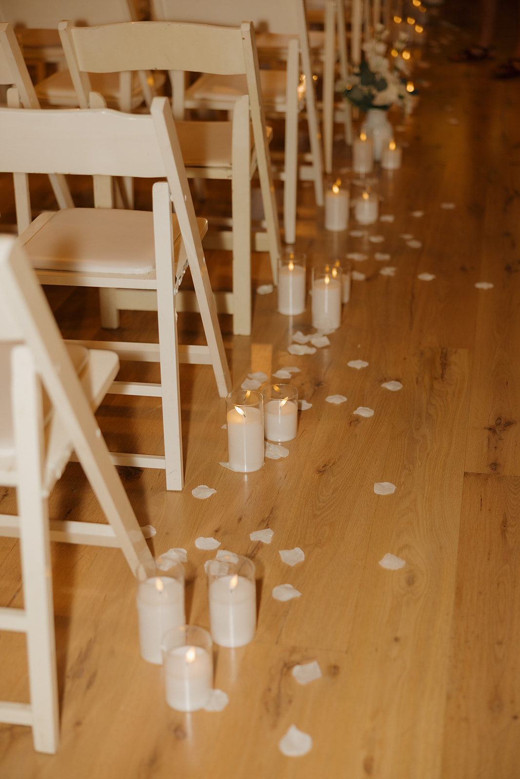 Soft white rose petals and glowing candles line the wooden aisle between white folding chairs, creating a romantic path to the altar.