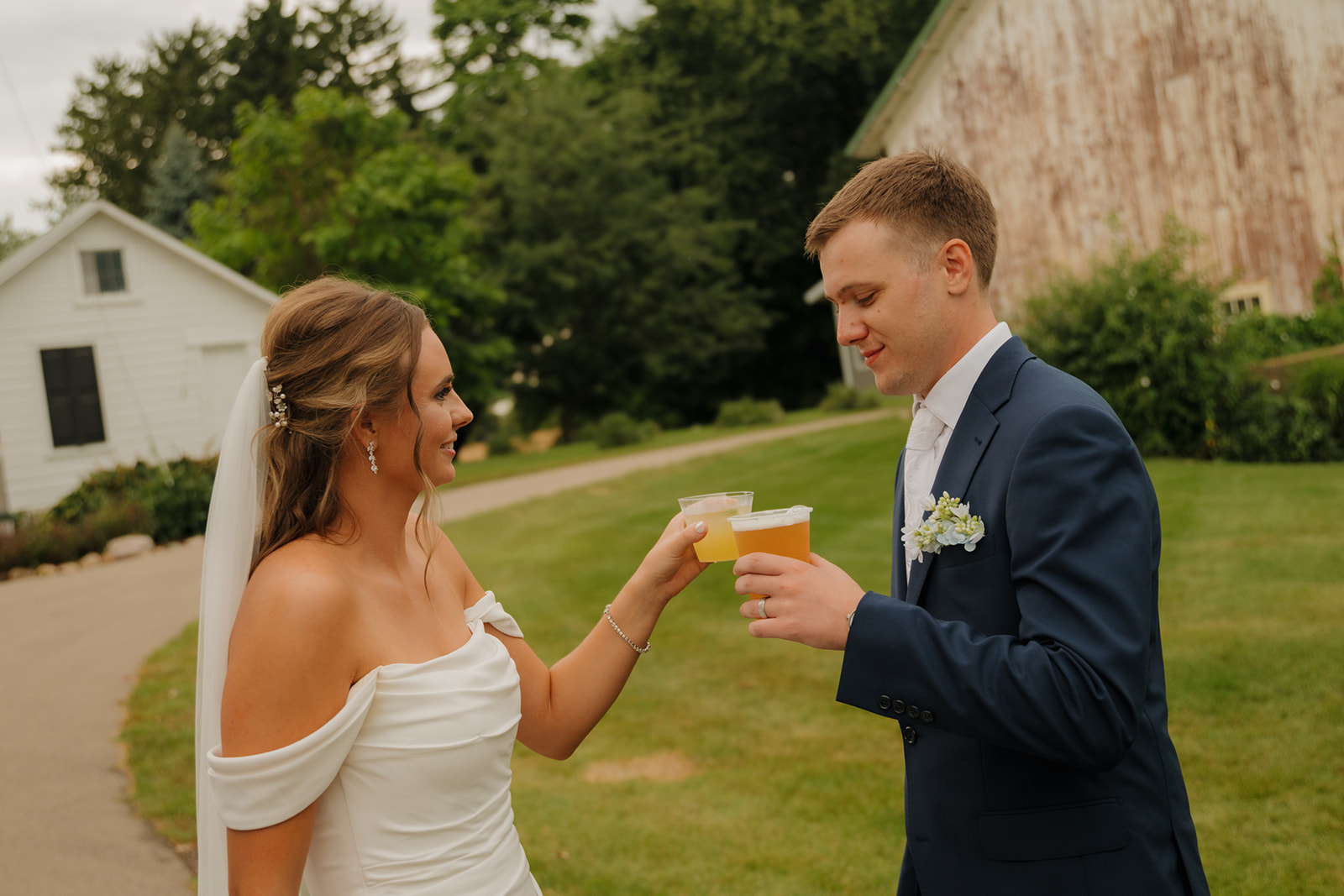 Newlyweds toast with beers outside a rustic venue, surrounded by greenery and soft overcast skies—an effortless moment documented by a wedding photographer in Wisconsin.