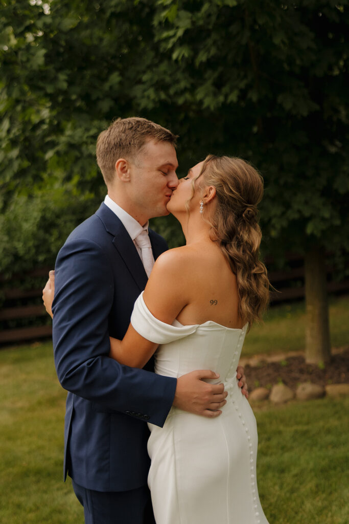 An intimate kiss between the bride and groom, the bride’s back tattoo peeking out as they hold each other close in a lush green setting.