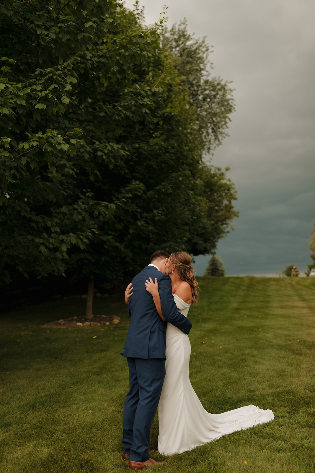 The couple shares an emotional embrace on a grassy hill, framed by moody clouds and lush trees—an intimate moment captured by a wedding photographer in Wisconsin.