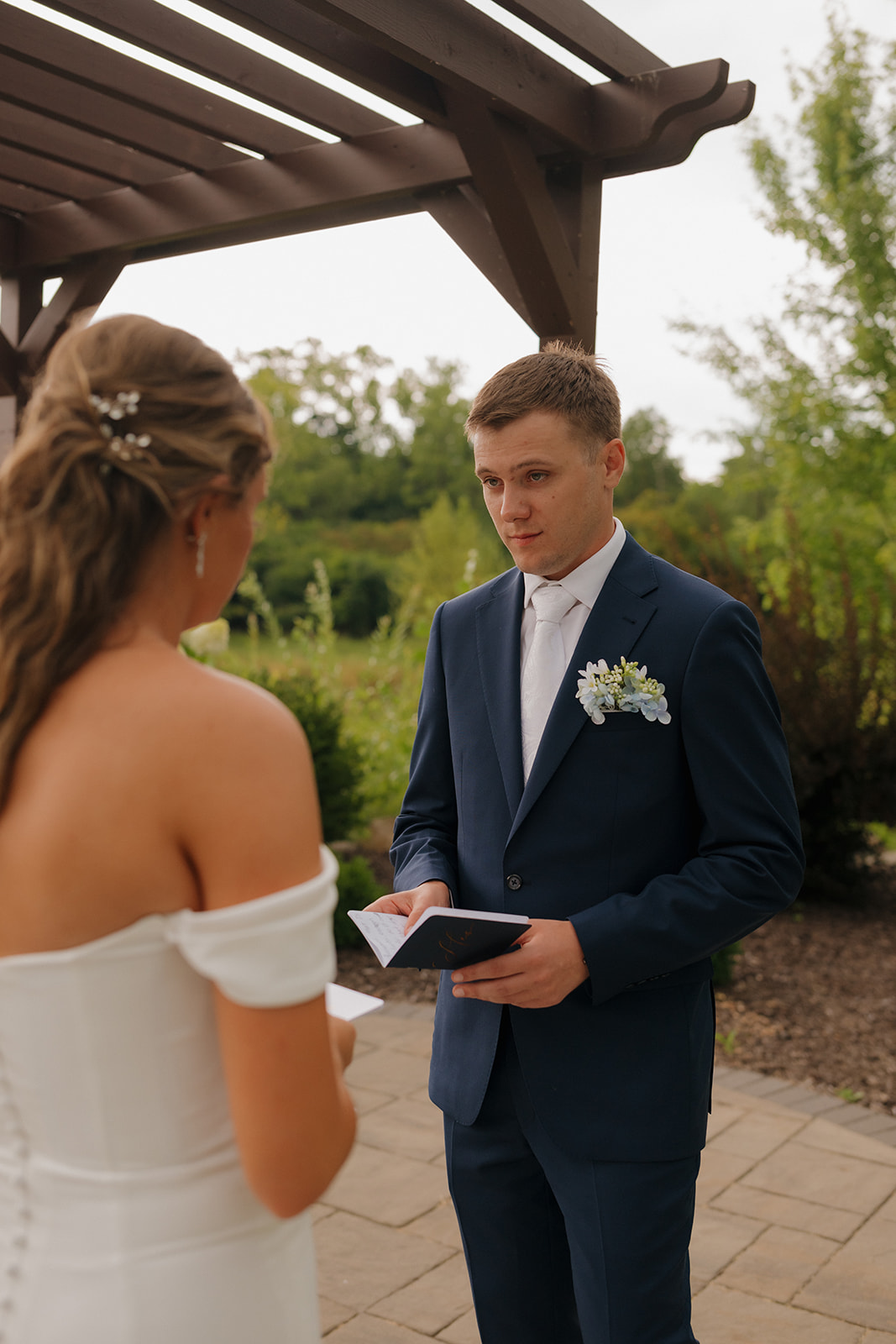 A groom reads handwritten vows to his bride during an intimate outdoor ceremony, standing beneath a wooden pergola on a cloudy day.