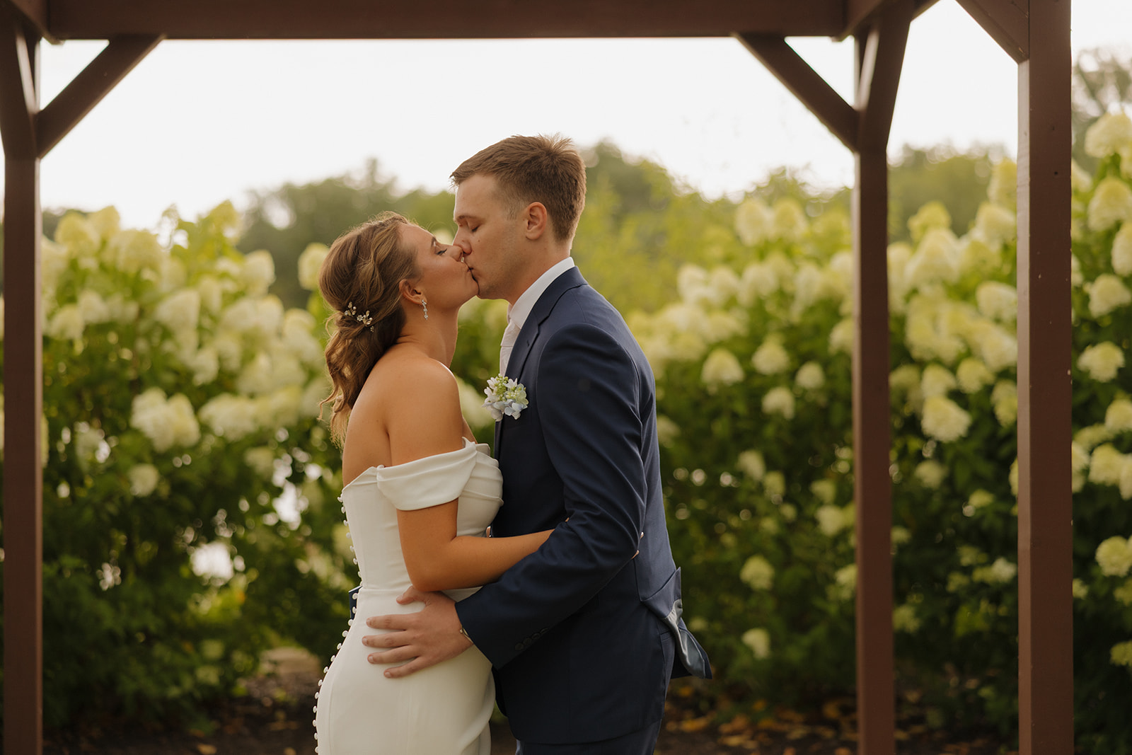 The couple shares a kiss under a wooden arbor surrounded by white hydrangeas, a sweet outdoor moment captured by a wedding photographer in Wisconsin.