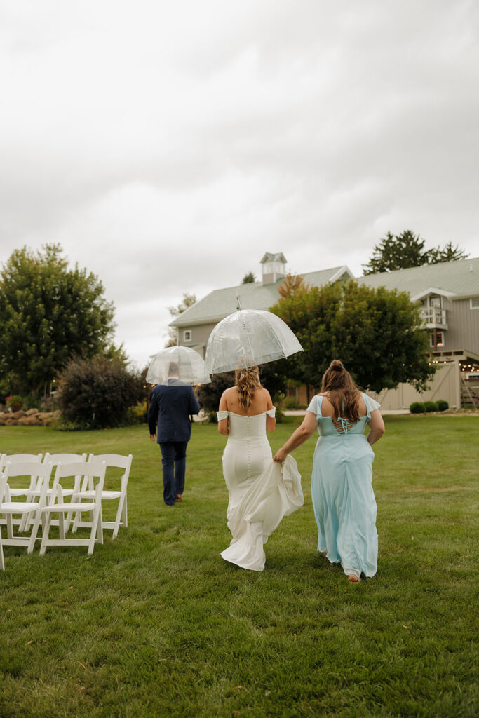The bride walks across the lawn under a clear umbrella, her dress held up by a bridesmaid, while gray skies hint at a romantic rainy-day wedding vibe.