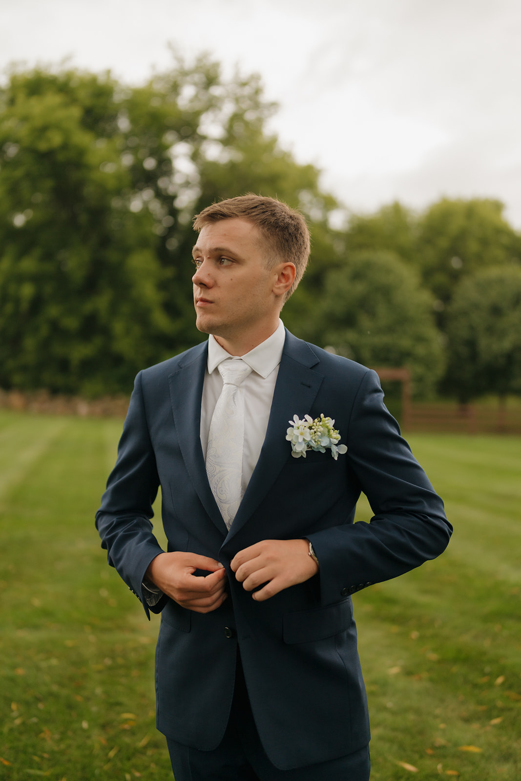 The groom stands in a grassy field adjusting his suit jacket, looking off into the distance with calm anticipation.
