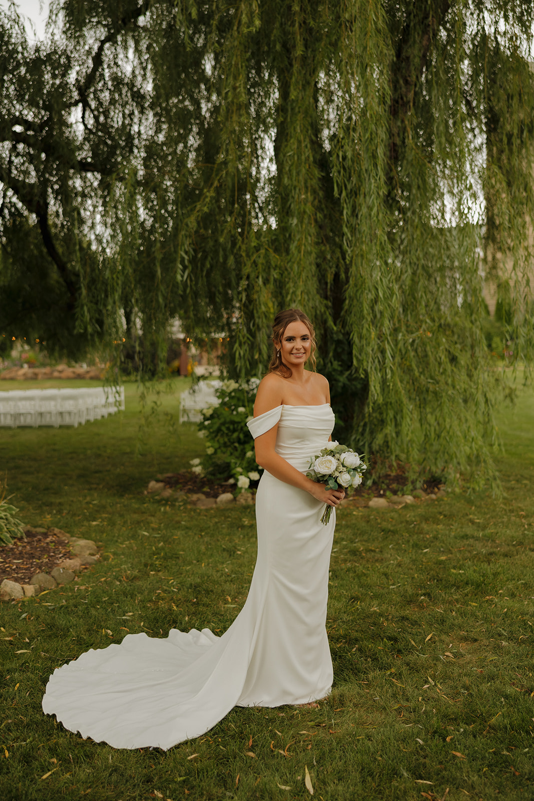 The bride poses in a fitted off-the-shoulder gown with a white bouquet under a sweeping willow tree, a serene setting for a wedding photographer in Wisconsin to document timeless beauty.