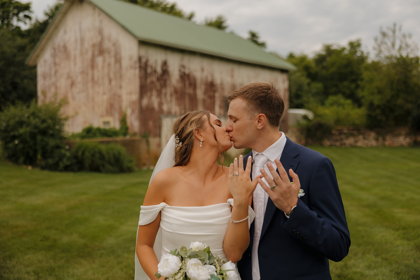 Bride and groom share a celebratory kiss while showing off their rings, standing in front of a weathered barn—classic charm captured by a wedding photographer in Wisconsin.