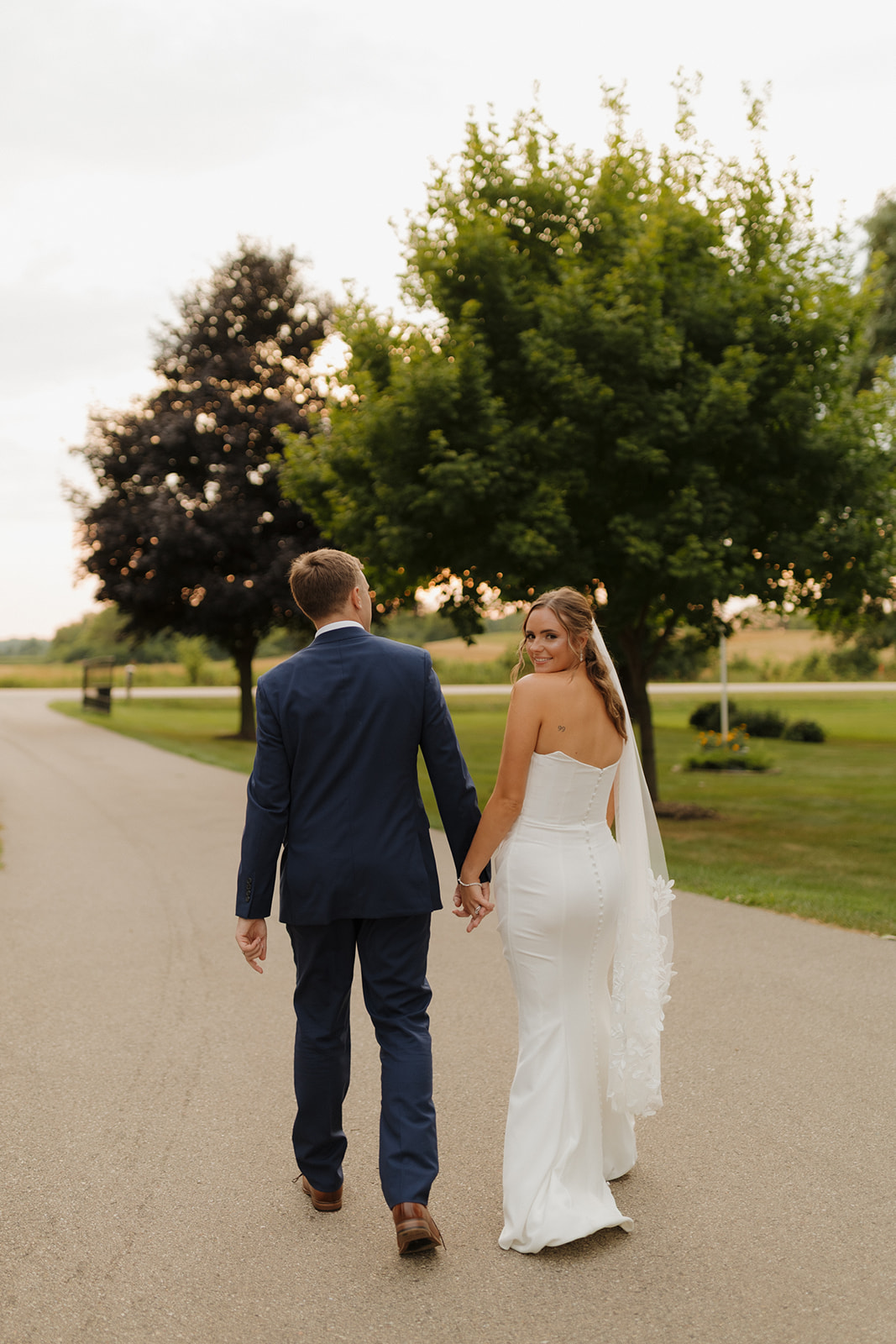 Newlyweds walk along a paved path, holding hands as the bride looks over her shoulder with a soft smile—Wisconsin farmland stretching out behind them.