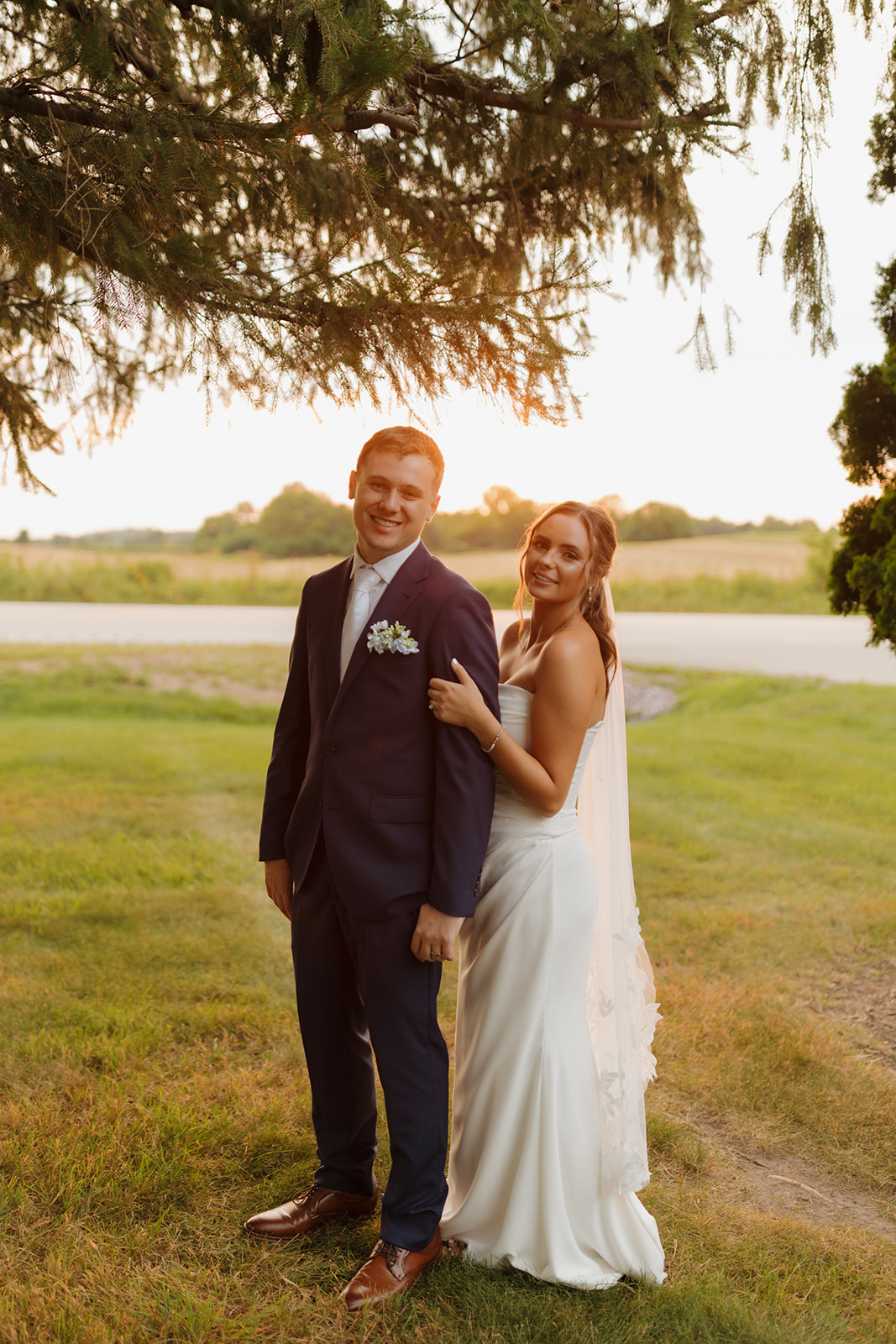 With warm evening light behind them, the bride and groom pose with relaxed smiles under a canopy of pine trees—an easygoing moment frozen in time by a wedding photographer in Wisconsin.