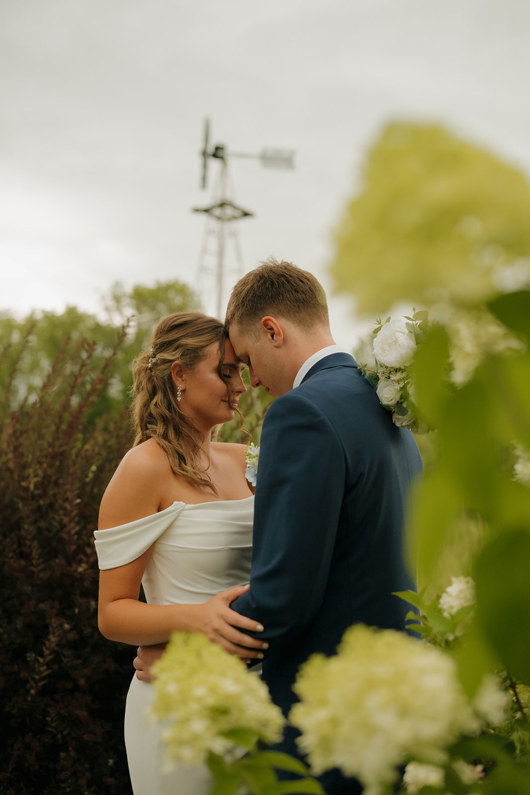 The couple shares a peaceful forehead-to-forehead moment surrounded by greenery and hydrangeas, with a windmill in the distance—captured by a wedding photographer in Wisconsin.