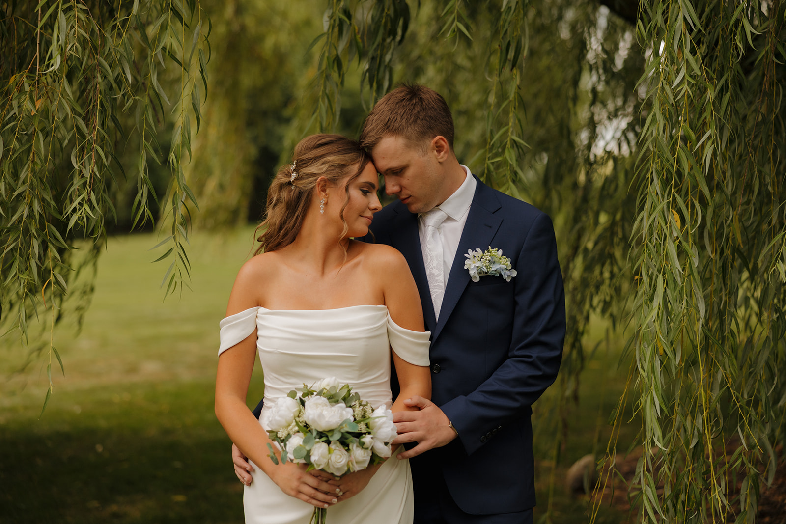 The couple shares a quiet, tender moment under a draping willow tree, their foreheads touching and the bride holding a bouquet of white flowers.