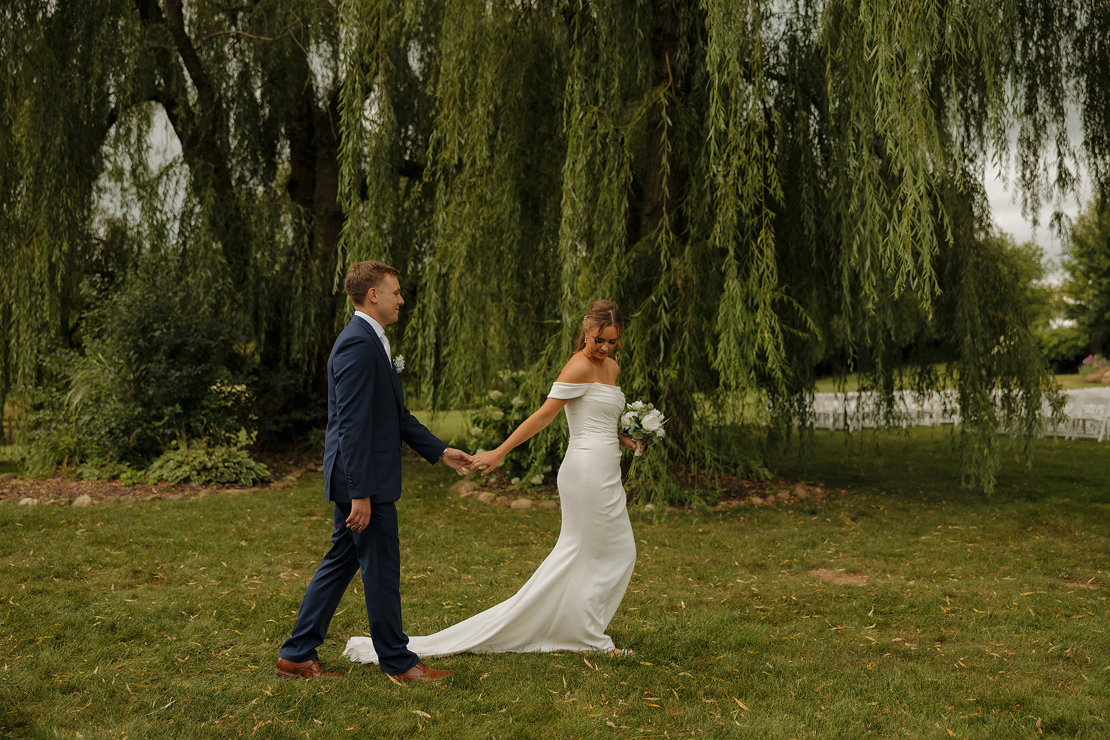 The newlyweds stroll hand-in-hand beneath a dramatic weeping willow, her train trailing behind and smiles soft—captured by a wedding photographer in Wisconsin.