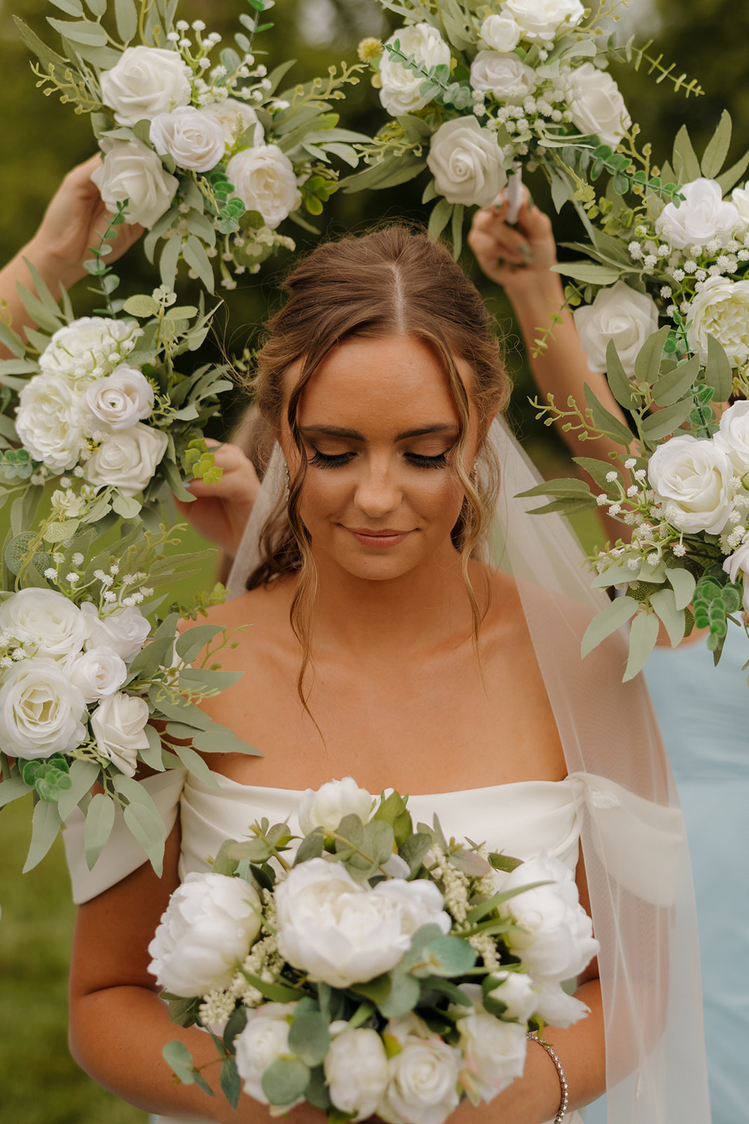 The bride looks down with a soft smile as her bridesmaids frame her with white floral bouquets, highlighting her serene beauty and elegant off-the-shoulder gown.