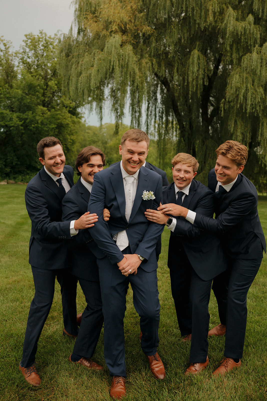 The groom shares a laugh with his groomsmen during a candid moment outdoors, all dressed in navy suits with brown shoes under a lush willow tree.