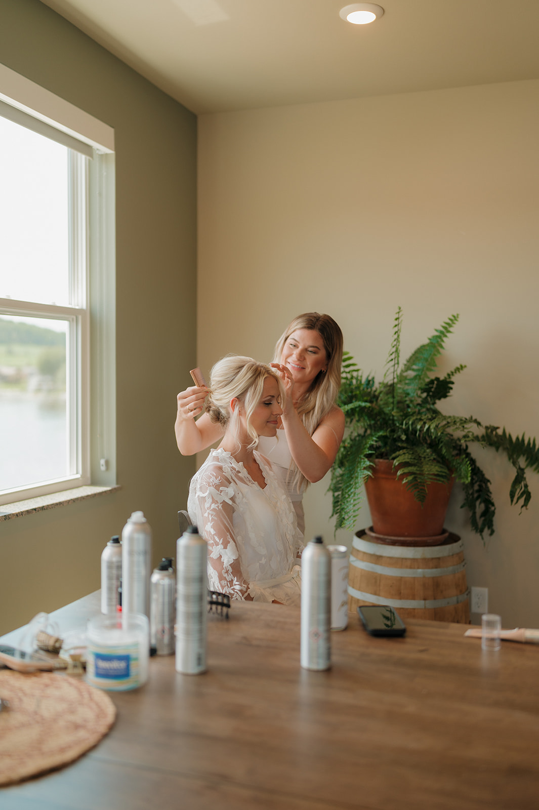 Bride getting ready by the lake with hairstylist and natural light before her winery wedding in Wisconsin.