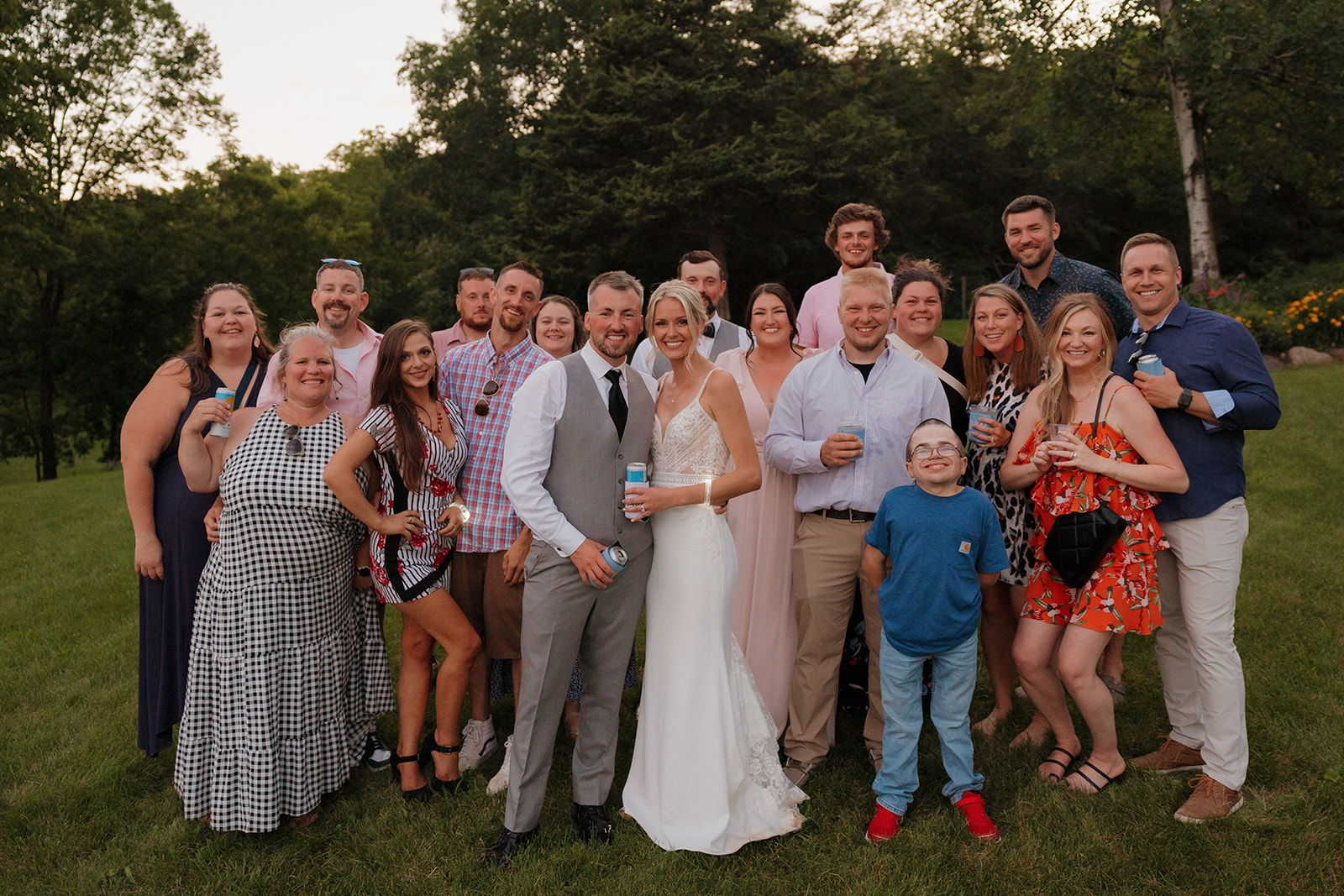 Bride and groom pose with a large group of smiling guests on a grassy hill.