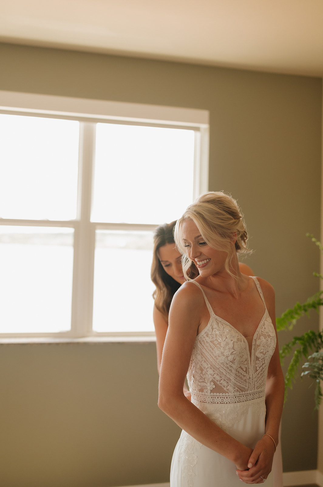 Bridesmaid laughs with the bride as they get ready by a sunlit window at a Wisconsin winery wedding venue.